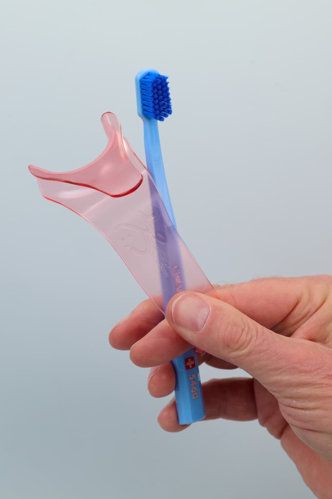 Eine Hand hält den Lippen- und Wangenhalter zusammen mit einer Curaprox CS 5460 Zahnbürste, die Bürste ist etwa so lang wie der Halter